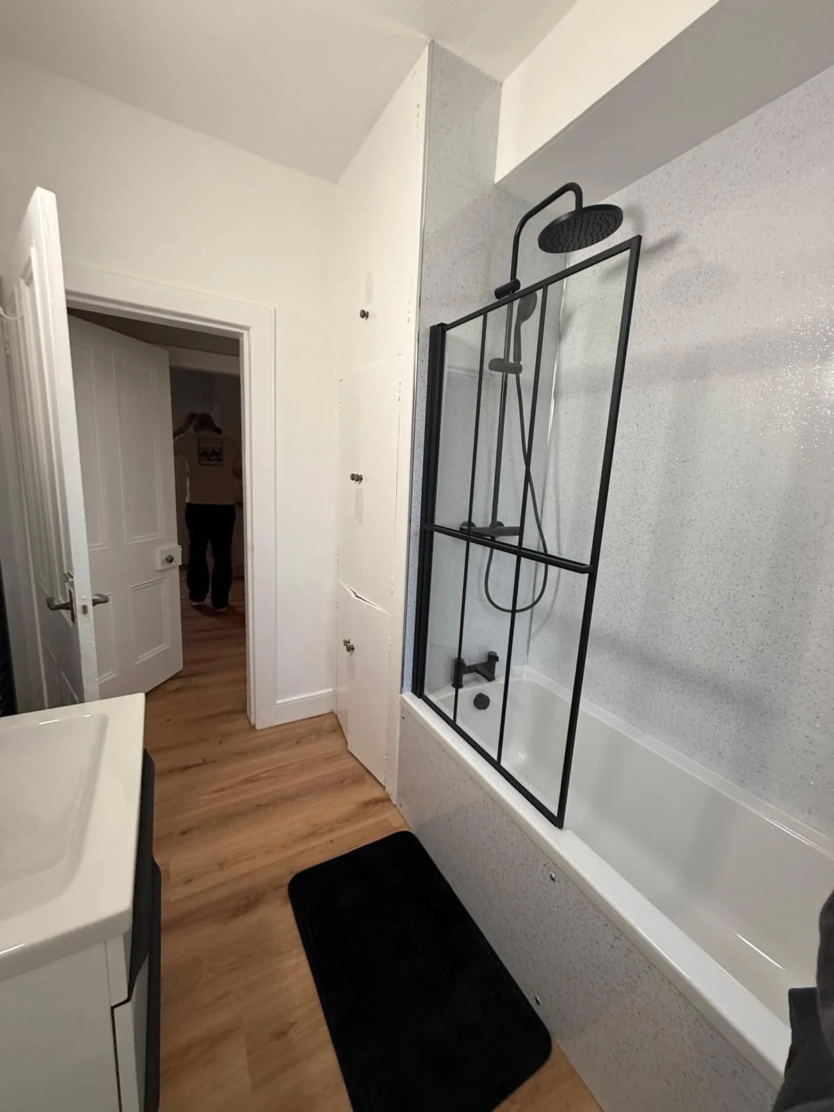 Bathroom with a bathtub, black-framed glass shower door, showerhead, a black bath mat, wood floors, white walls, and a partially open door leading to another room.