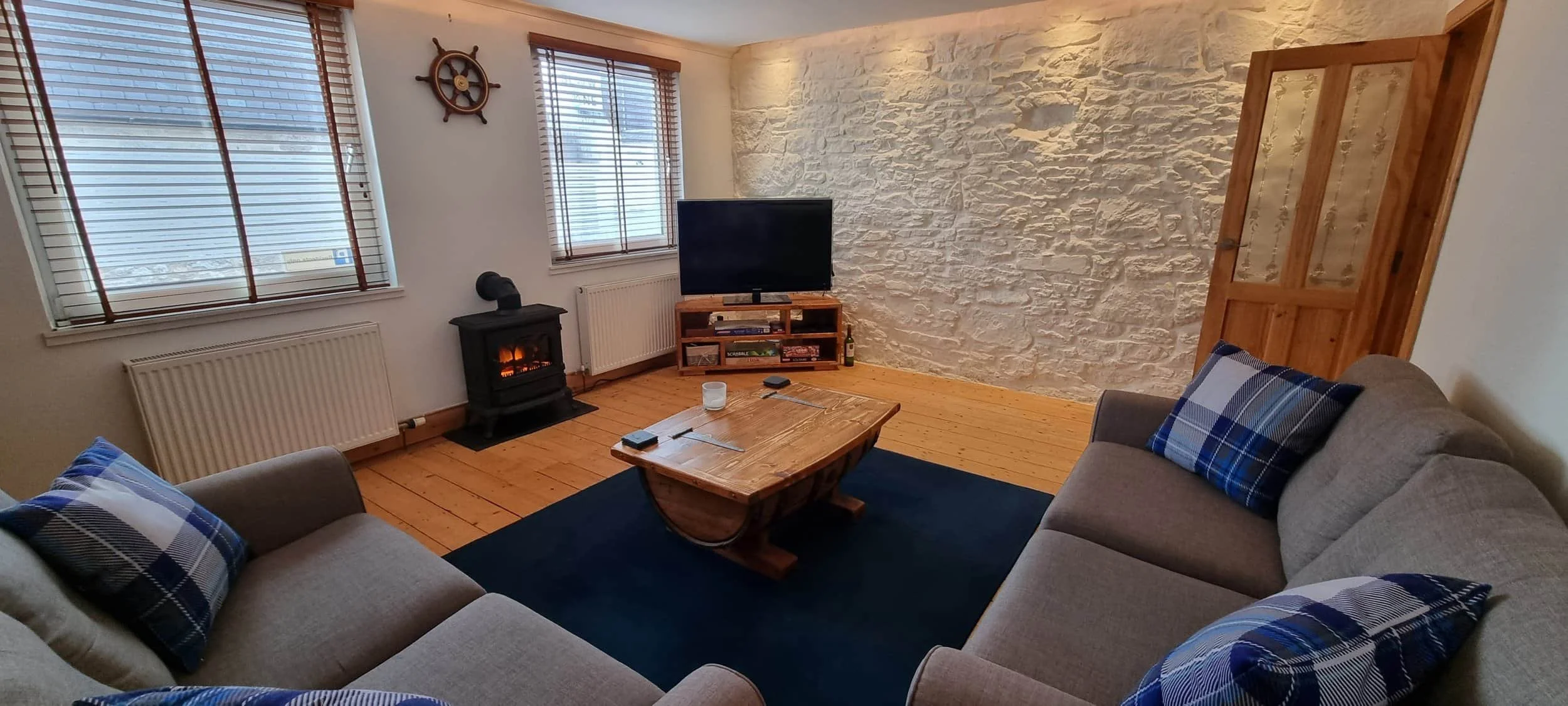 Living room with two beige sofas with blue plaid pillows, a wooden coffee table, a black electric fireplace, a flat-screen TV on a wooden stand, a white stone accent wall, two windows with blinds, a wooden door, and a dark blue rug.