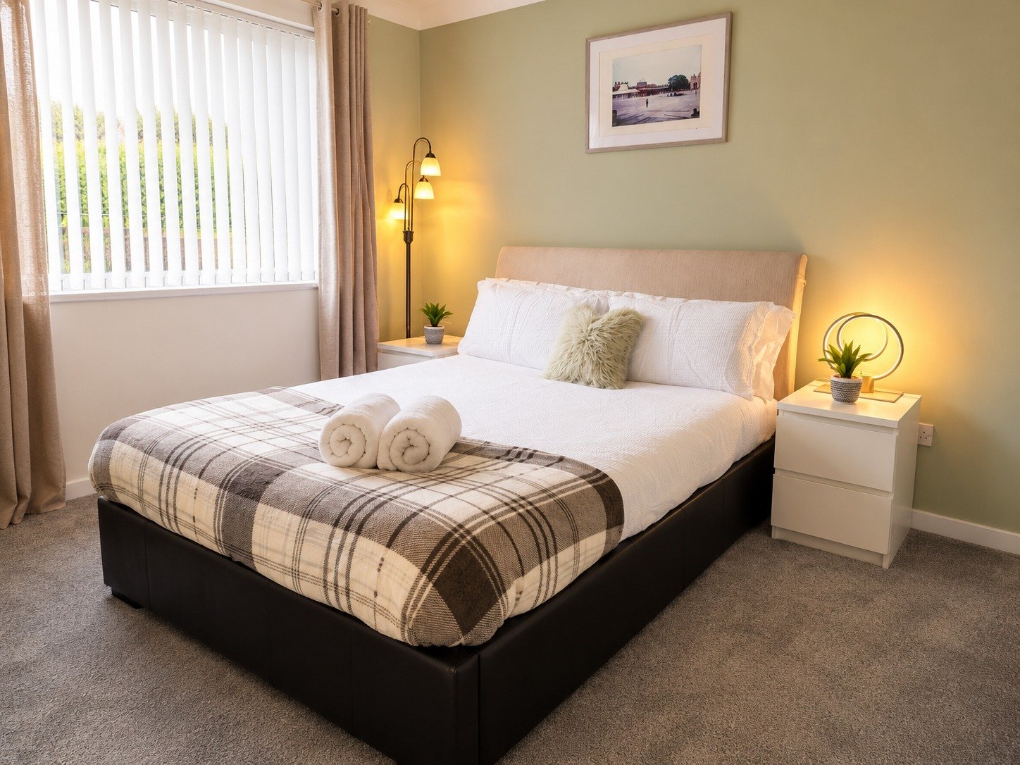 A neatly made bed with white linens, a plaid blanket at the foot, and two rolled towels on top. The bed has a beige headboard and a green accent pillow. There are two white nightstands with small potted plants and decorative lamps on either side of t