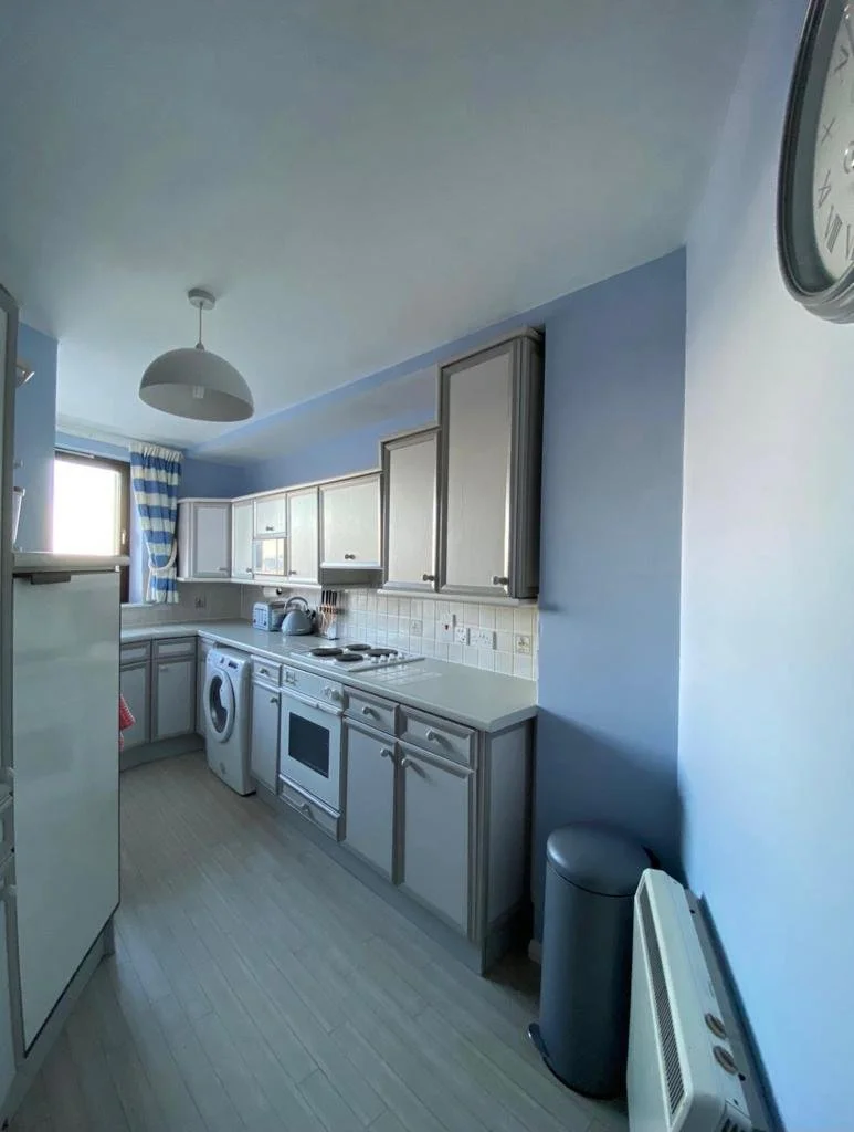 Small kitchen with blue walls, white cabinets, a washing machine, oven, kettle, and a window with blue and white striped curtains.