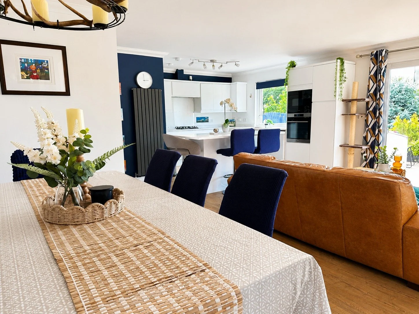 Open-concept dining and kitchen area with a table set with a woven runner and a flower arrangement, a tan leather sofa, and white cabinets in the background, with view to an outdoor patio.