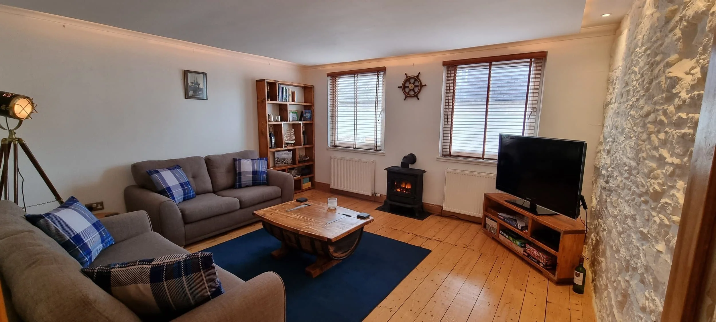Living room with two gray sofas with blue plaid pillows, wooden coffee table, blue rug, wood flooring, bookshelf, window blinds, decorative ship wheel, electric fireplace, flat-screen TV on wooden stand, stone accent wall, and a bottle of wine.