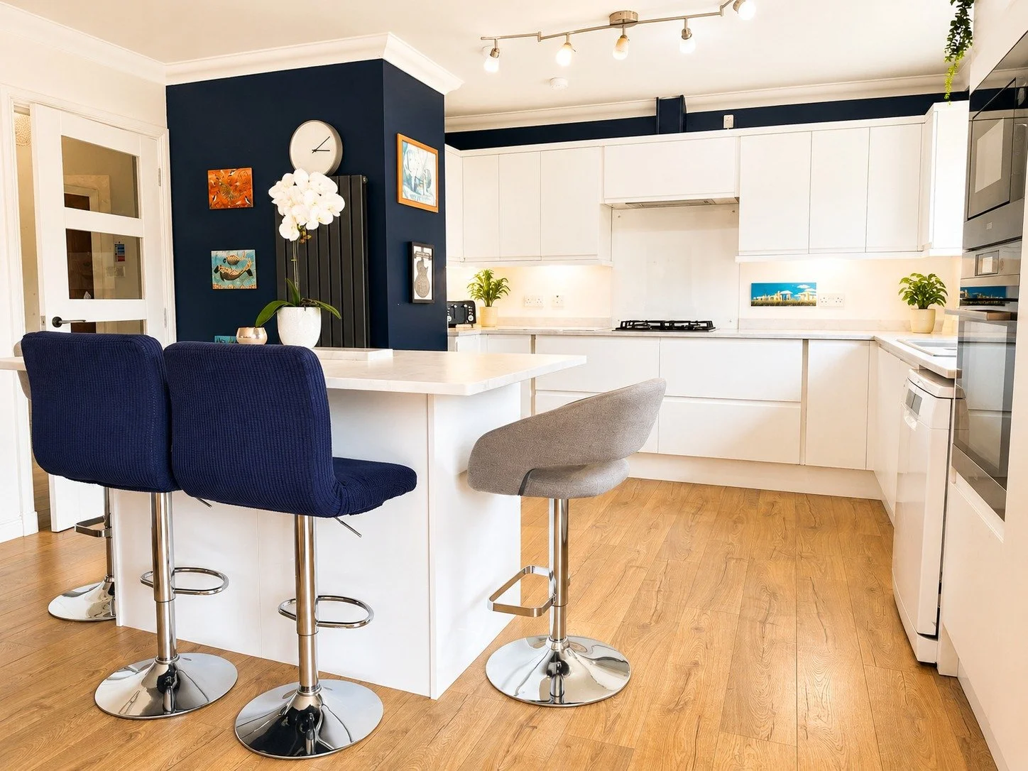 Modern kitchen with white cabinets, a white island with seating, and a dark blue accent wall decorated with art and a clock. There are two navy blue barstools and one grey barstool around the island. The room has light wood flooring and some potted p