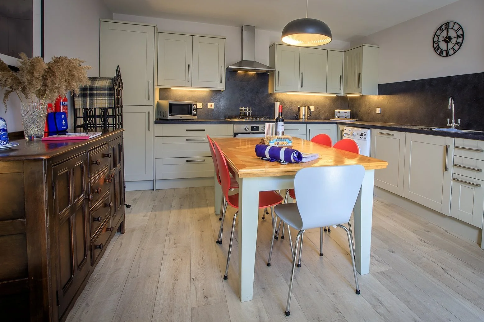 Modern kitchen with white cabinets, black countertops, a wooden dining table with red and white chairs, and various appliances including a microwave, kettle, and dishwasher, decorated with a clock on the wall.