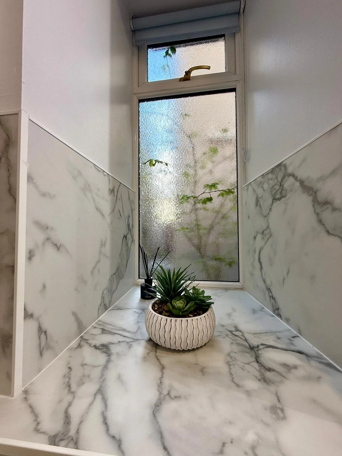 A small windowsill with a white ceramic pot containing green succulents, a black reed diffuser, and a window with frosted glass and a small brass handle, surrounded by marble-like wall and countertop surfaces.