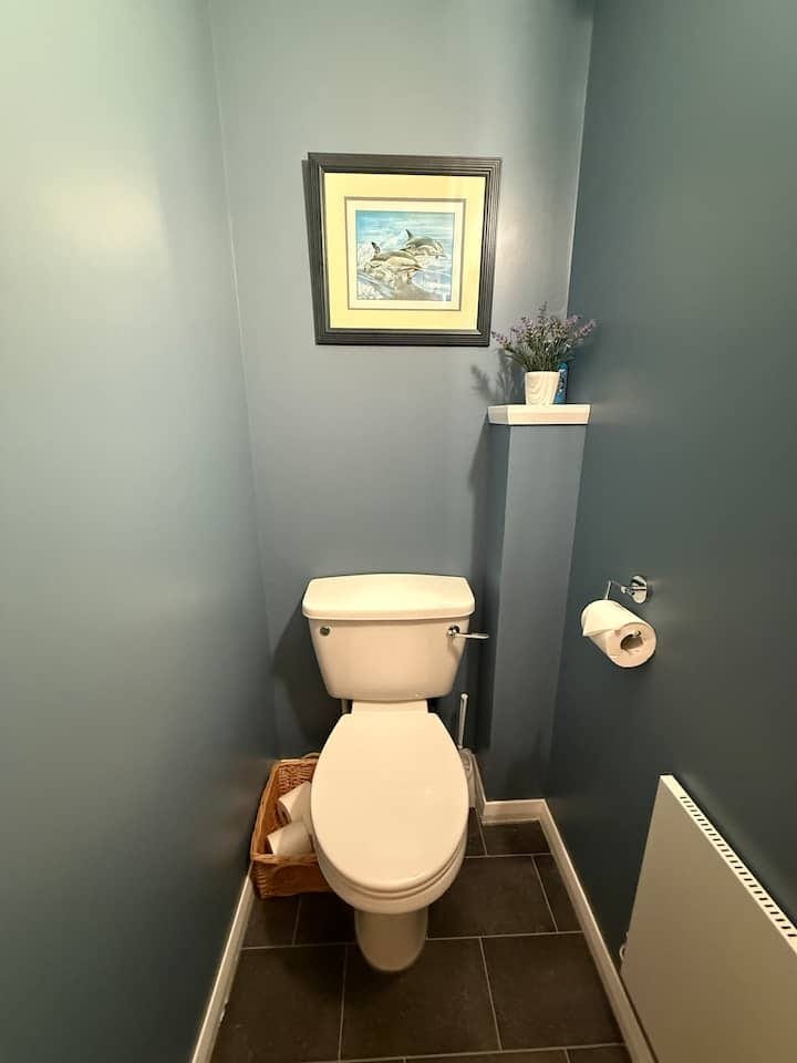Small bathroom with a white toilet, wicker basket with toilet paper on the floor, a framed picture of dolphins above the toilet, a shelf with purple flowers above the toilet, a toilet paper roll on the wall, and a radiator heater on the right.