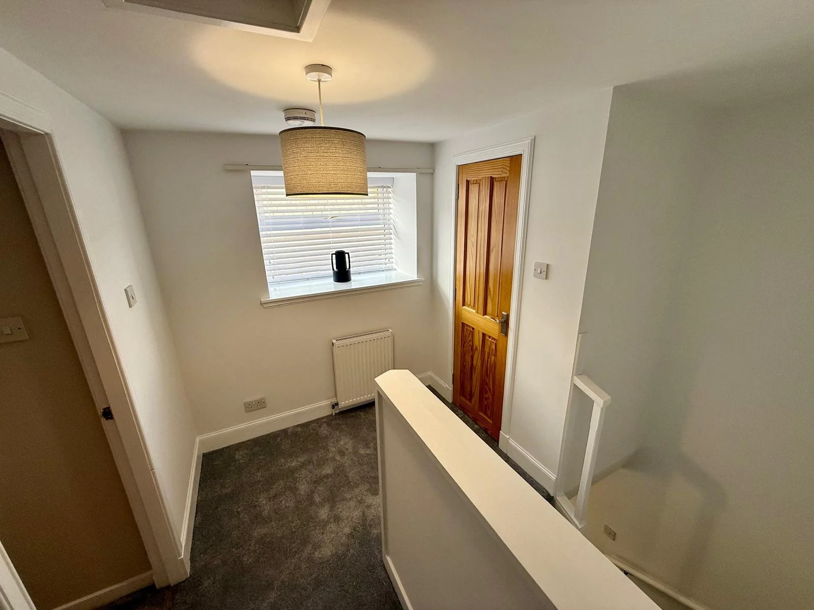 Furnished small hallway with white walls, a window with blinds, a wooden door, a gray carpet, a ceiling light with a drum shade, a radiator below the window, and a staircase leading down to a lower level.