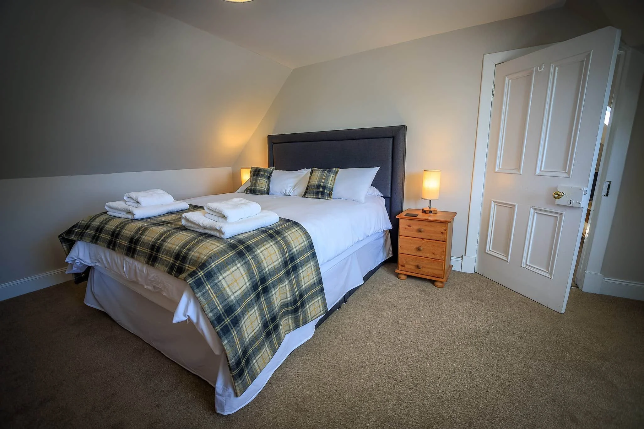 A neatly made bed with white linens and a plaid bed runner in yellow, black, and green, topped with three pillows, two with matching plaid covers and one plain white. On the bed are three folded white towels. There is a wooden nightstand with a small