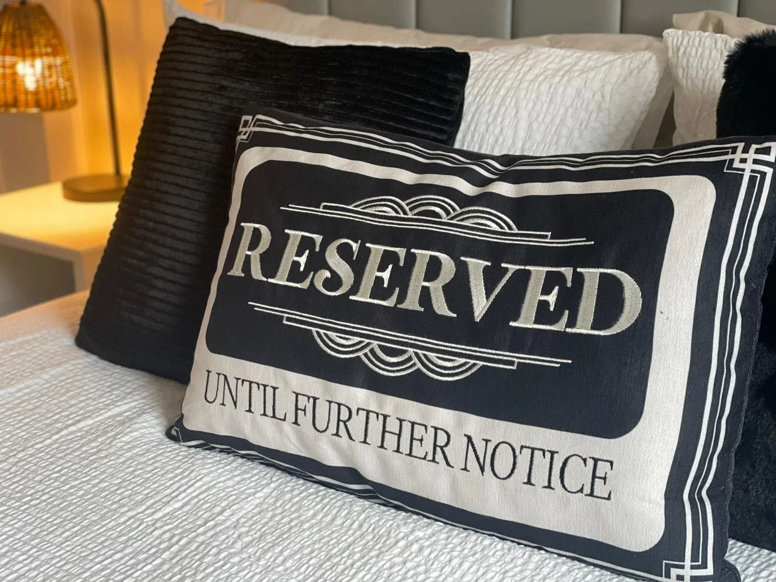 A bed with three pillows, one with a reserved sign and the other two plain black and white, in a room with a nightstand and a lamp in the background.