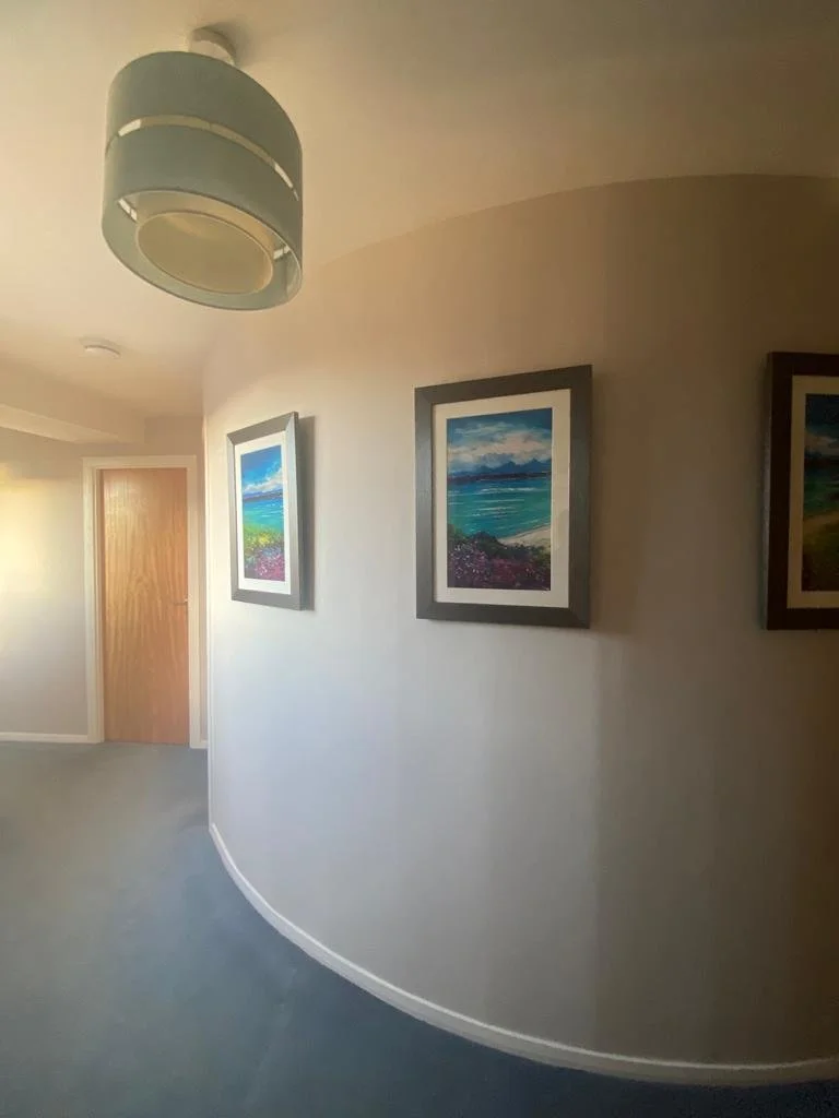 Hallway with three landscape paintings on the wall, a wooden door at the end, beige walls, a gray carpet, and a modern ceiling light fixture.