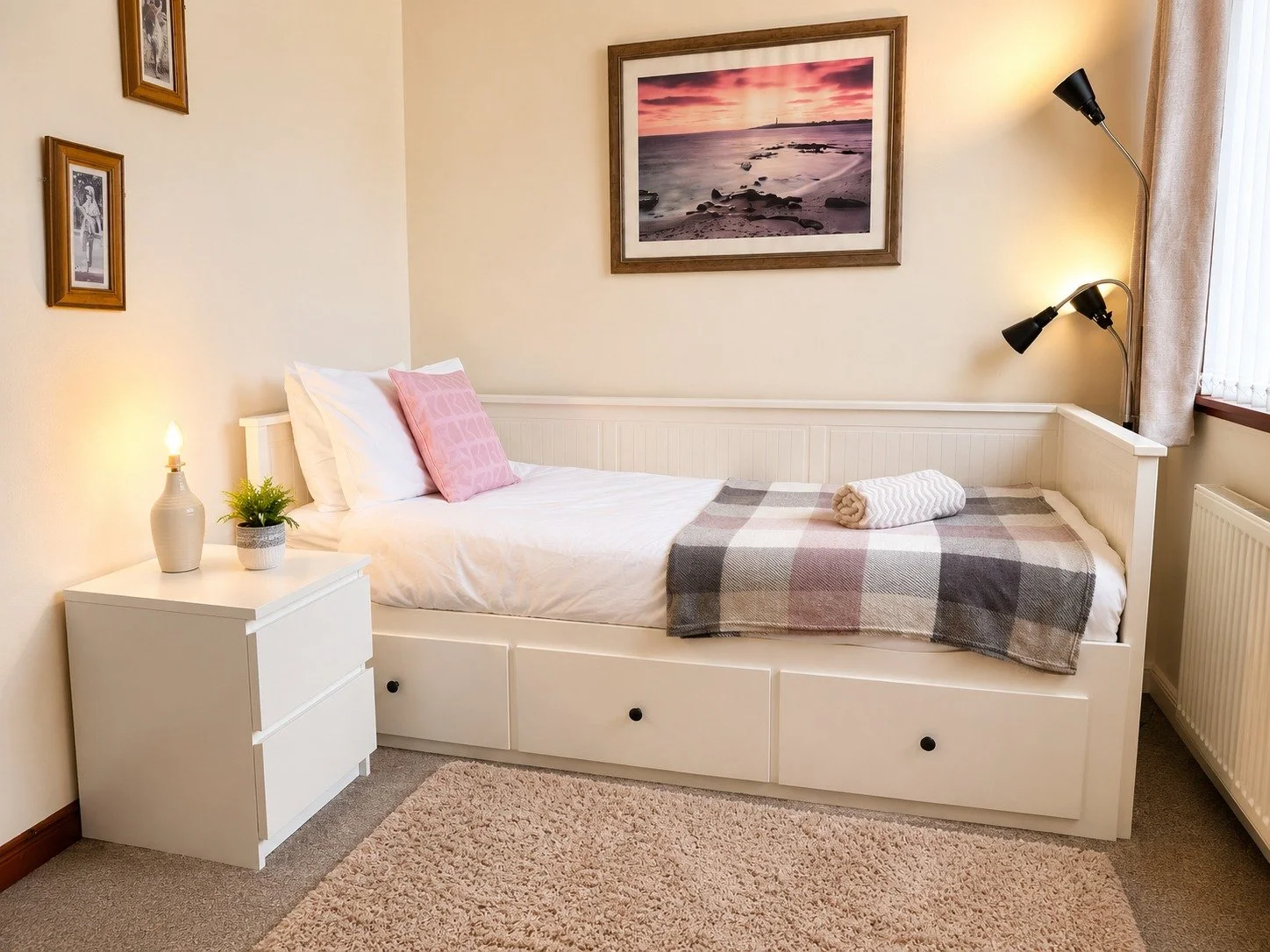A cozy bedroom with a single bed, white frame, pink pillow, checkered blanket, rolled towel, nightstand with lamp and plant, wall art depicting a sunset over the beach, and a window with curtains.
