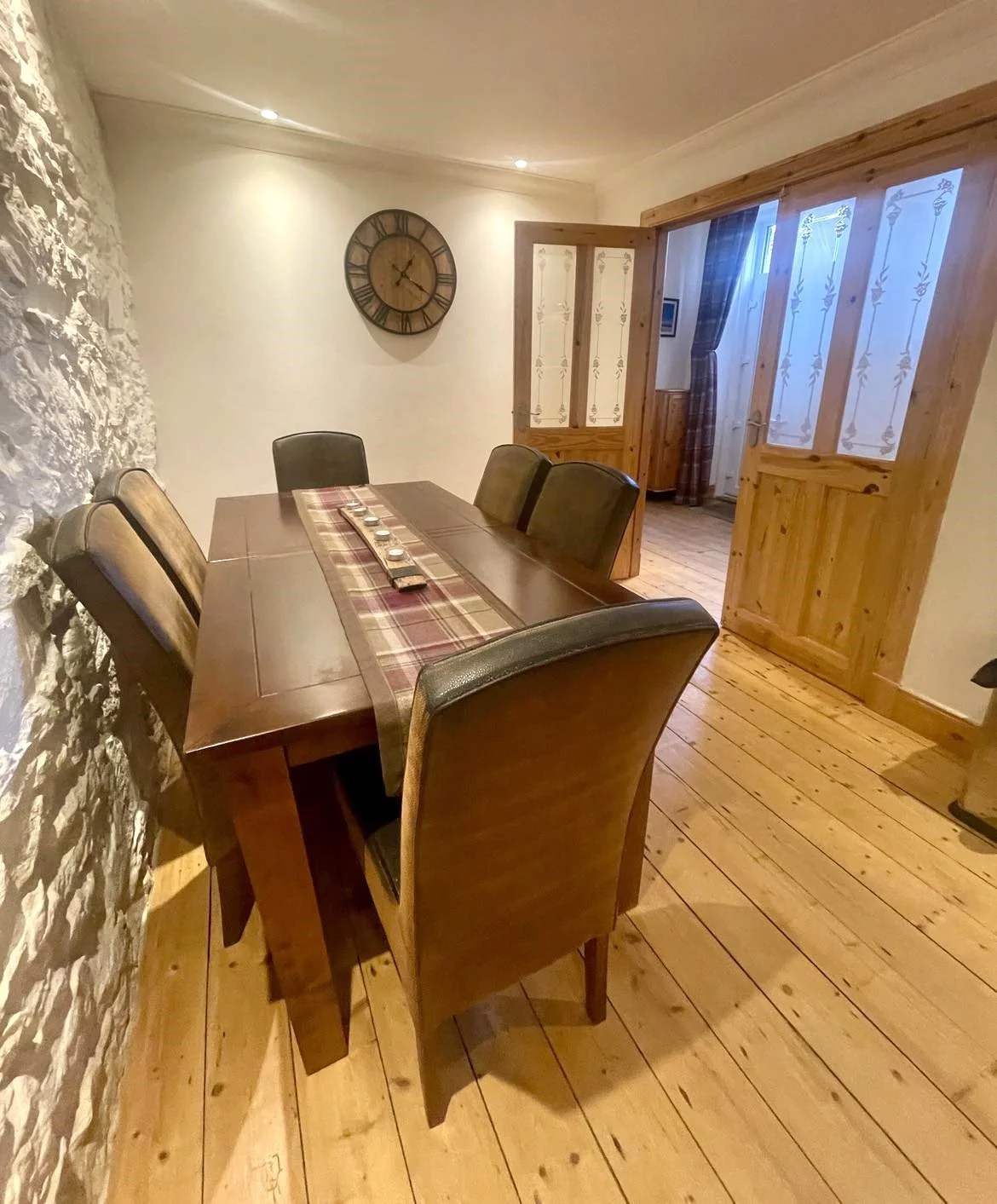 A dining room with a wooden table, six upholstered chairs, a stone wall on the left, a large wall clock, wooden sliding doors with frosted glass, and hardwood flooring.