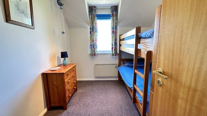 Small bedroom with a wooden bunk bed, a window with floral curtains, a small wooden dresser with a blue lamp, and from the door, a view into the room.