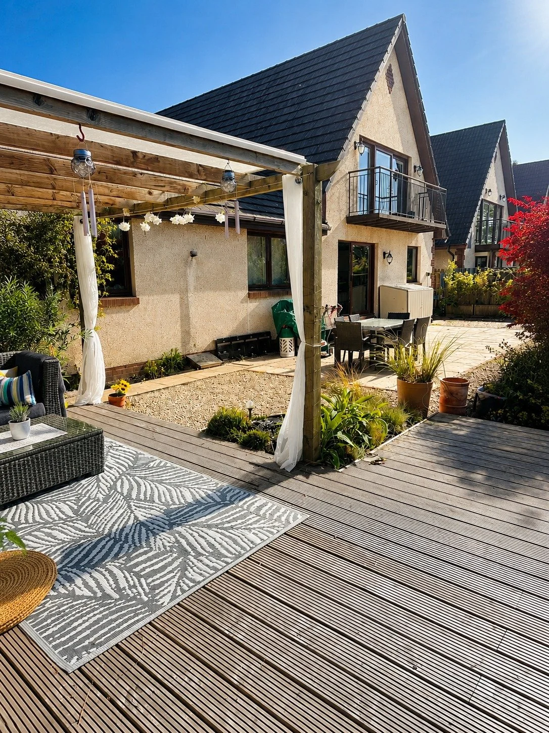 Backyard patio area with wooden decking, outdoor seating, potted plants, garden decoration, and a house with dark roof and balcony in the background.
