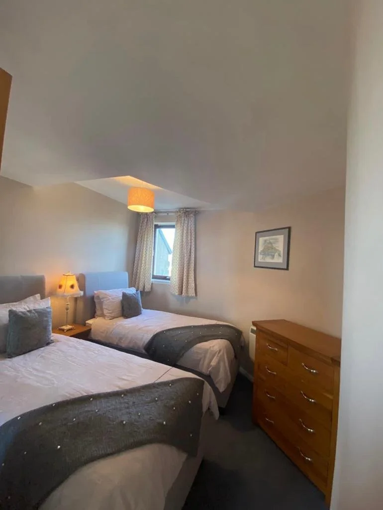 A bedroom with two beds, a window with curtains, a wooden dresser, a wall picture, and bedside lamps.