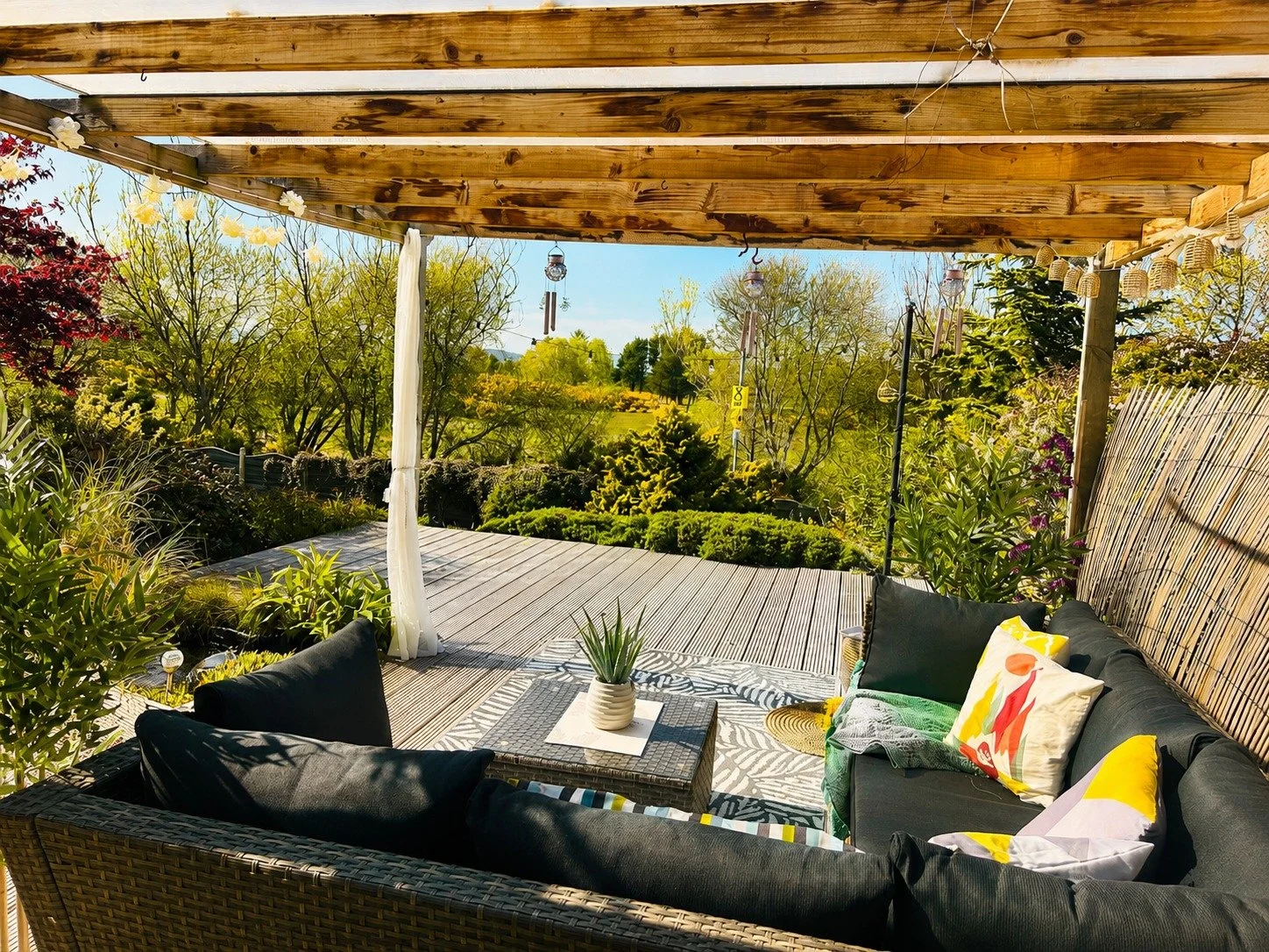 Outdoor patio with a black cushioned wicker sofa, cushions, a small wicker table with a plant, surrounded by lush green garden with trees, shrubs, and hanging wind chimes, under a wooden pergola on a sunny day.