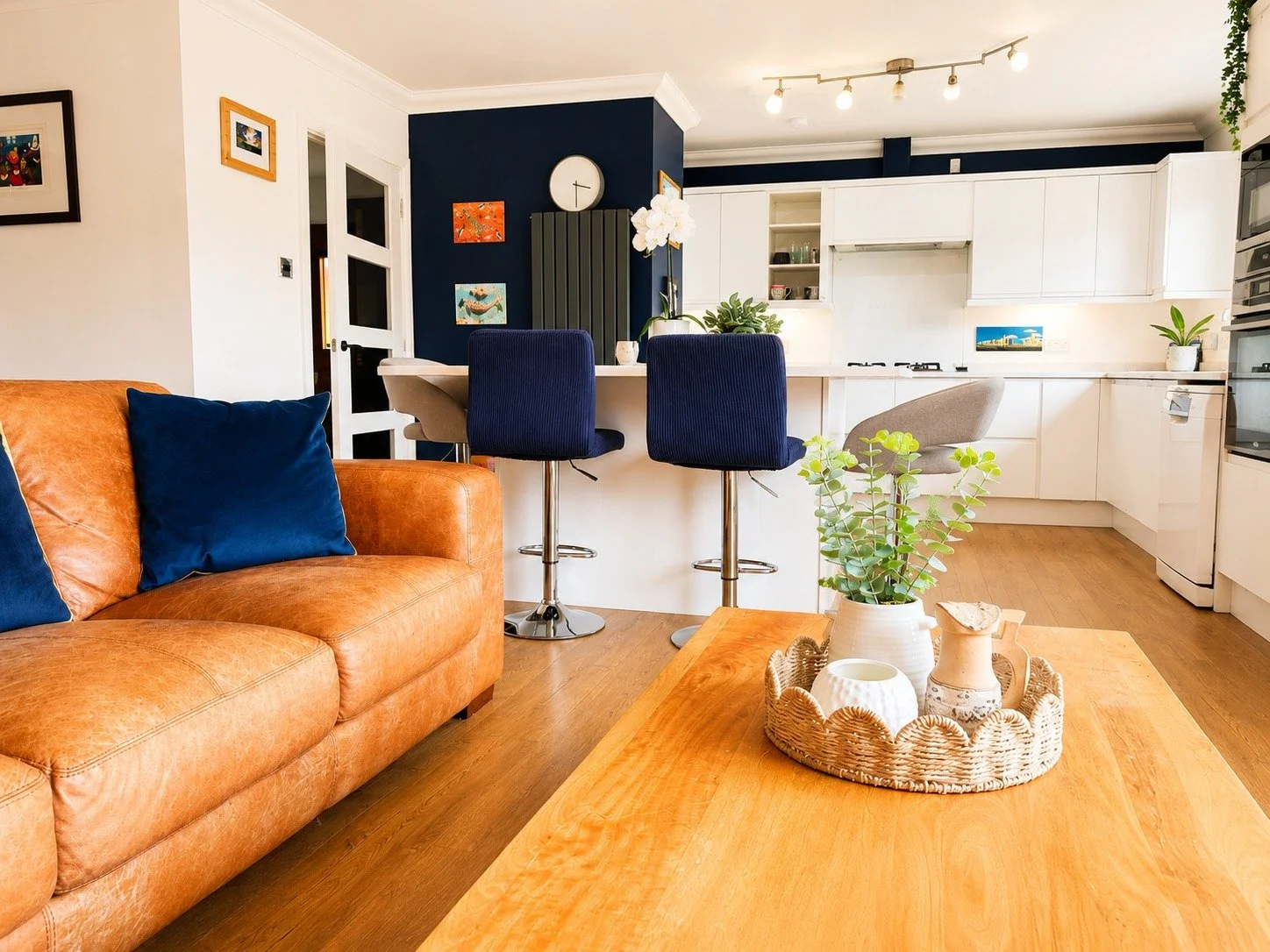 Living room with tan leather sofa with blue pillows, wooden coffee table with a plant and decorative items, open kitchen with white cabinets, blue accent wall, bar stools, and various wall art and plants.
