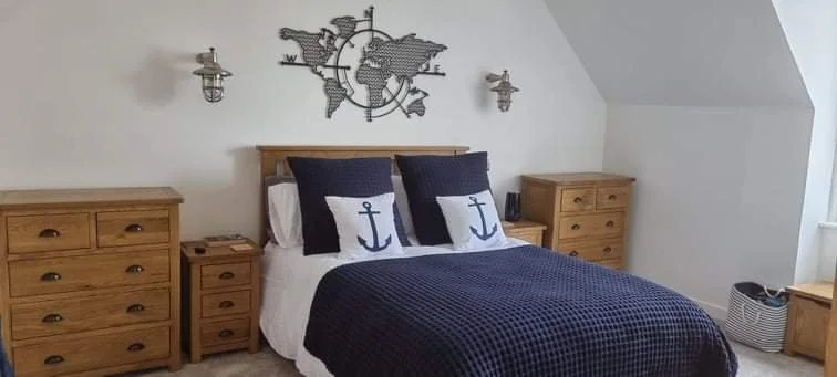 A cozy bedroom with a wooden bed frame, navy blue and white bedding with anchor pillows, two matching wooden nightstands, wall-mounted nautical-themed metal world map decor, and two wall sconces. There's a window on the right side.