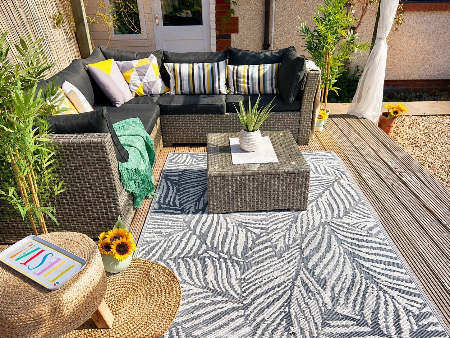 Outdoor patio with wicker furniture, including a black cushioned sectional sofa and a matching armchair, on a wooden deck. There are patterned pillows on the sofa, a small wicker coffee table with a potted plant, and a rug with a leaf design. Bright 