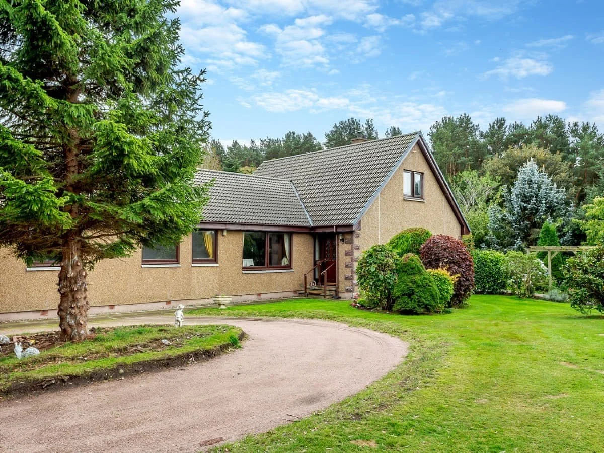 A house with beige walls and a brown tiled roof, surrounded by a lush green garden with various bushes, trees, and a curved gravel driveway, under a partly cloudy sky.