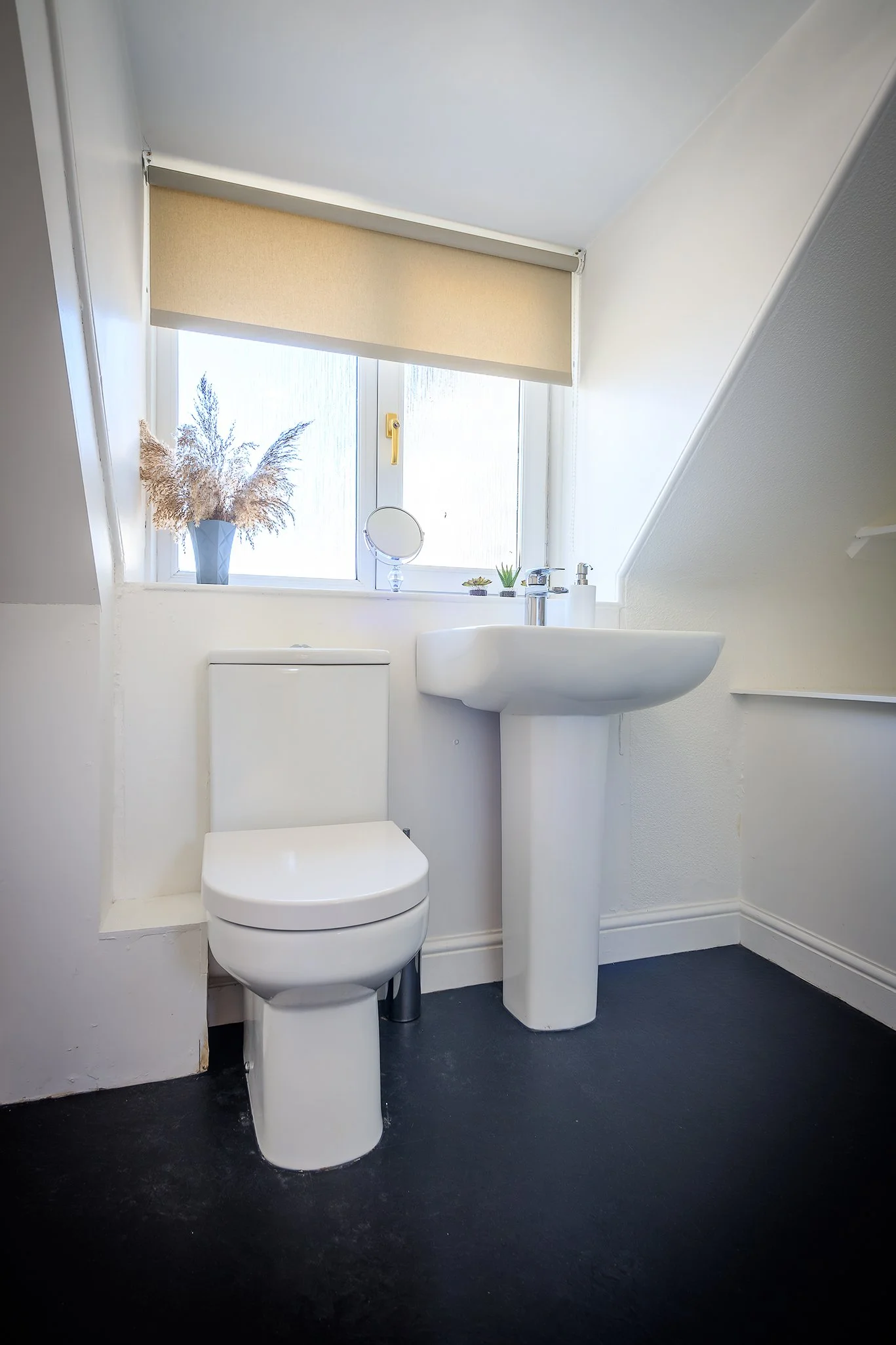 A small, clean bathroom with a white toilet and a pedestal sink, a window with a beige blind, and decorative items like a small round mirror, potted plants, and a vase of dried flowers on the window sill.