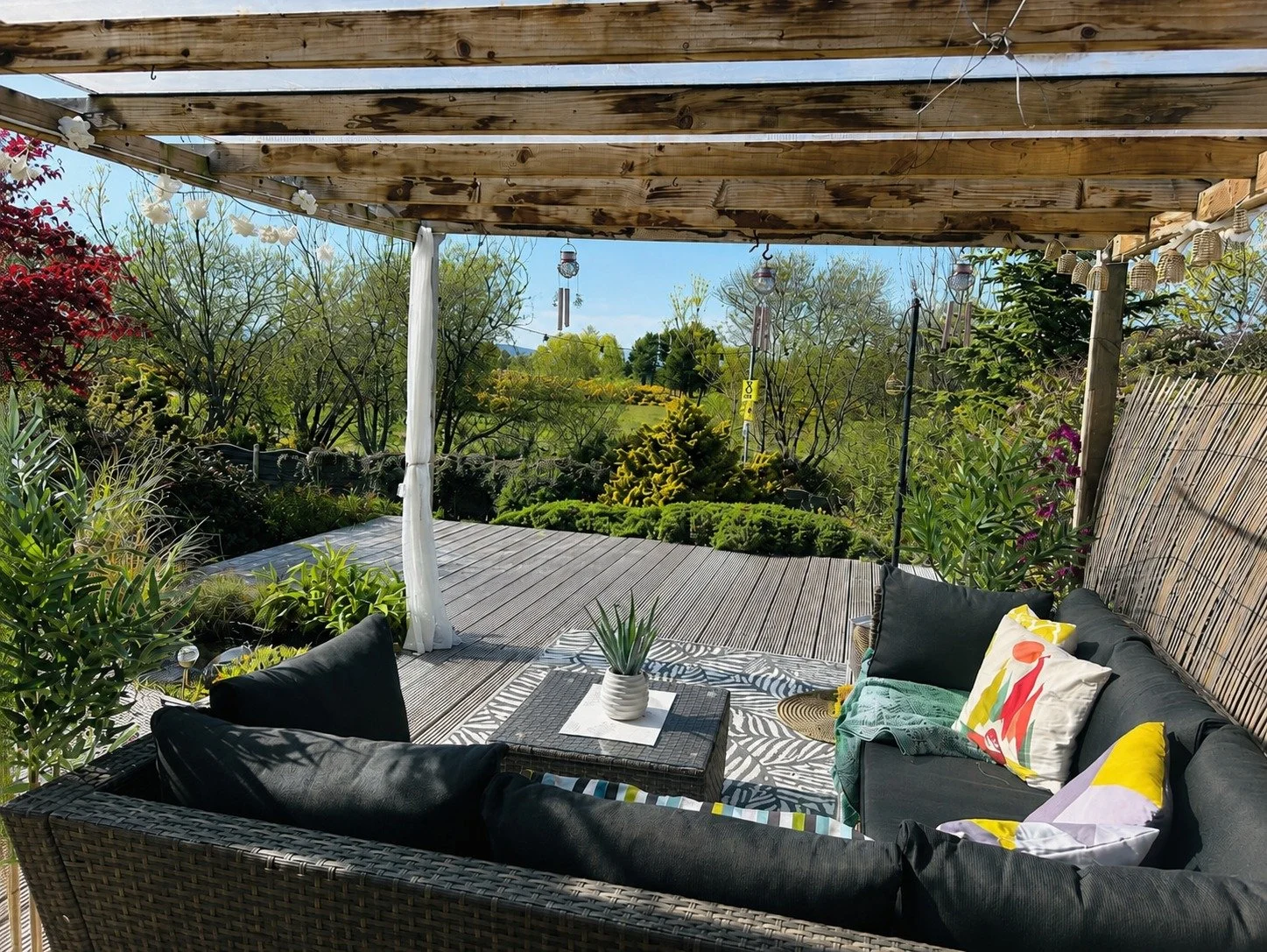 A cozy outdoor patio with black cushioned wicker furniture, some with colorful pillows, on a patterned outdoor rug. There's a small woven table with a potted plant in the center. The patio has a wooden pergola with string lights and decorative wind c