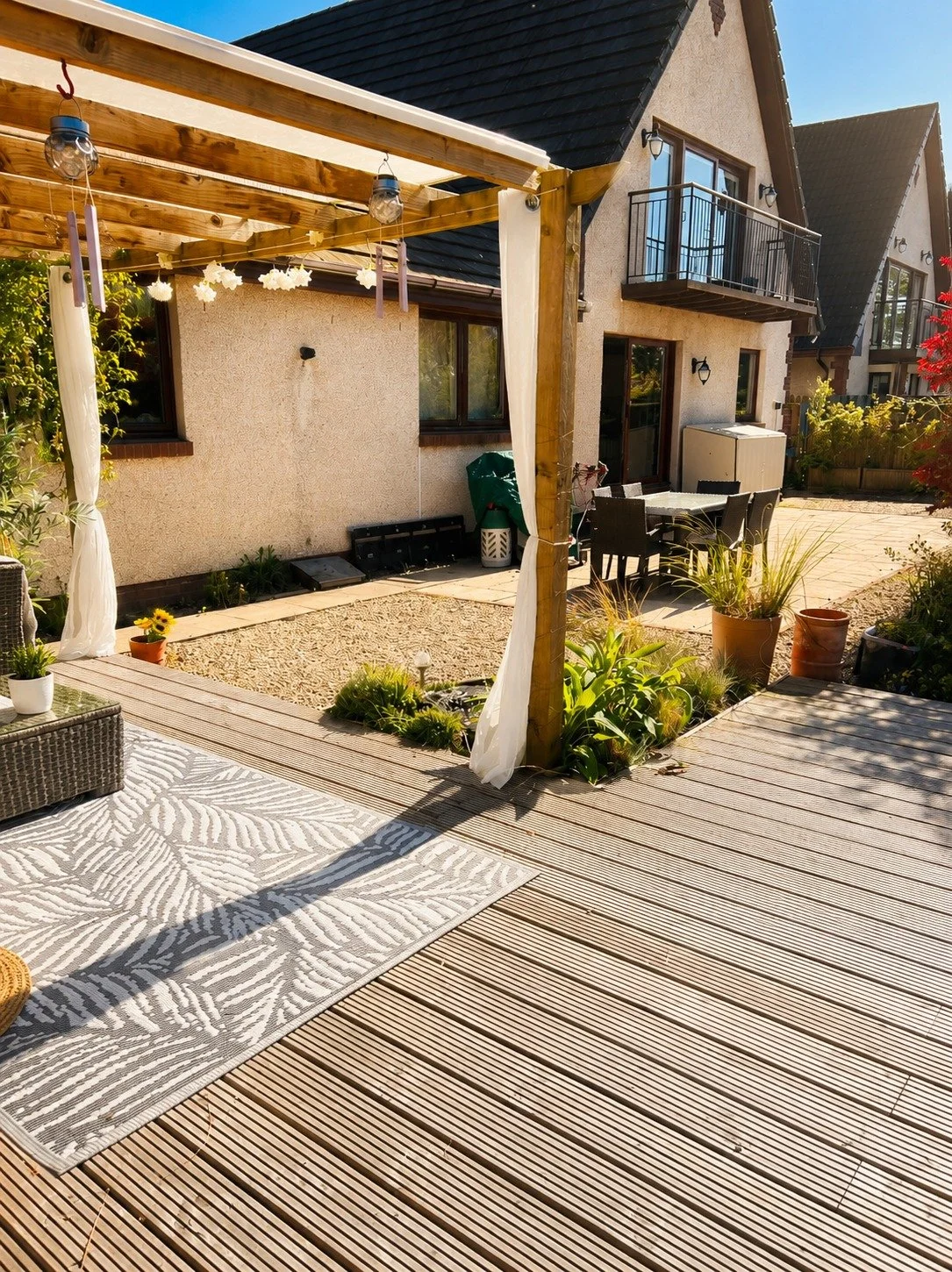 Backyard patio with outdoor seating, potted plants, and a wooden pergola in front of a house with balcony-style windows.