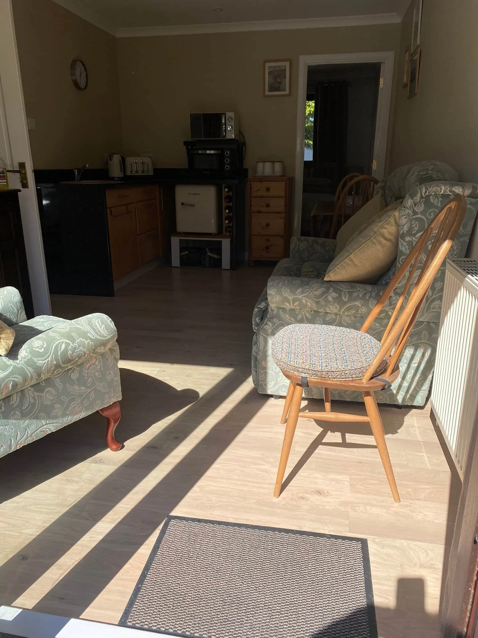Living room with sunlight casting shadows, featuring a patterned sofa with pillows, a wooden chair with a cushioned seat, a small doormat, and an open kitchen area with appliances and a doorway leading to another room.