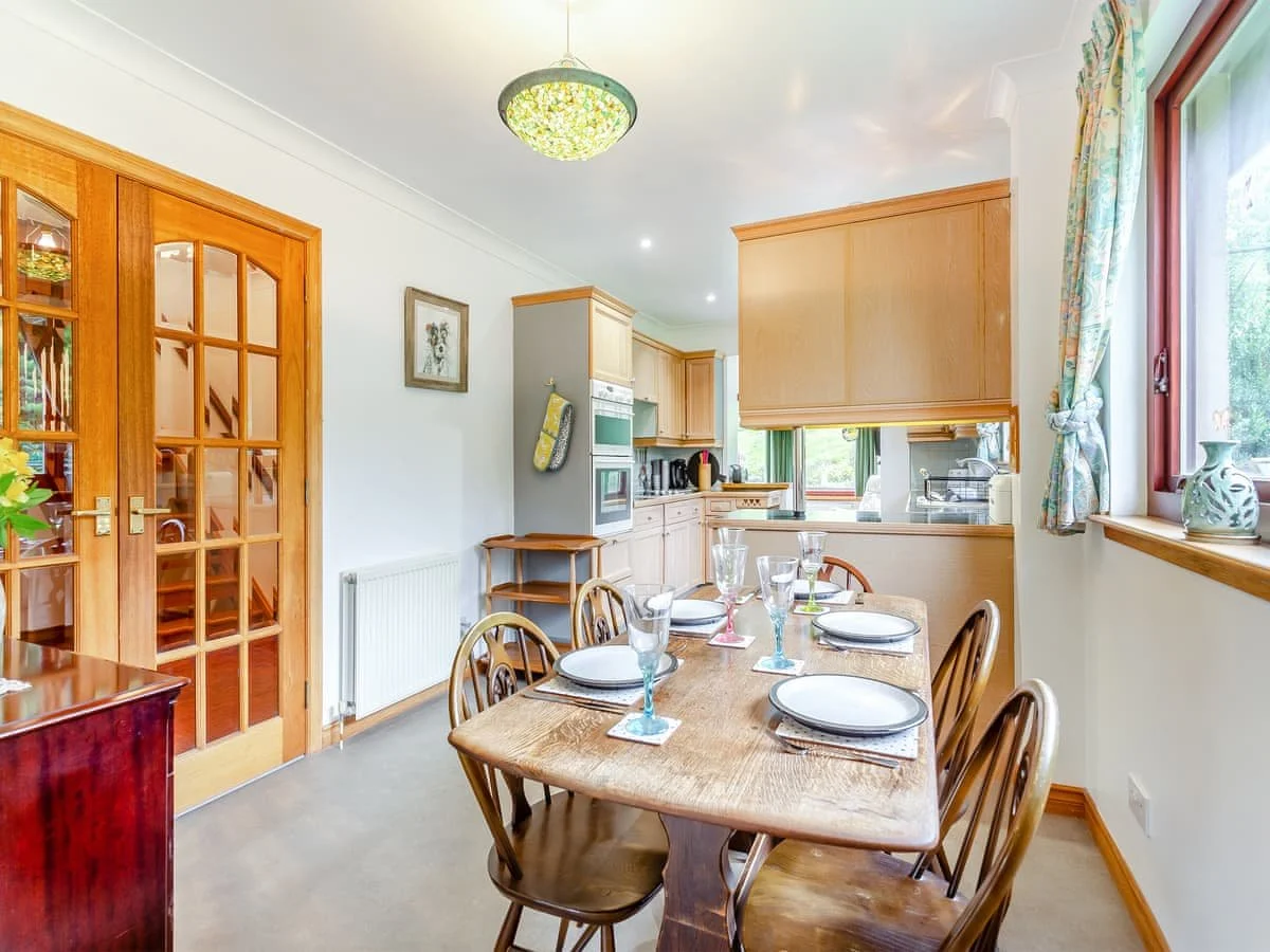 Dining area with a wooden table set with six plates, glasses, and utensils, surrounded by wooden chairs, adjacent to a kitchen with light wood cabinets and a window with floral curtains.