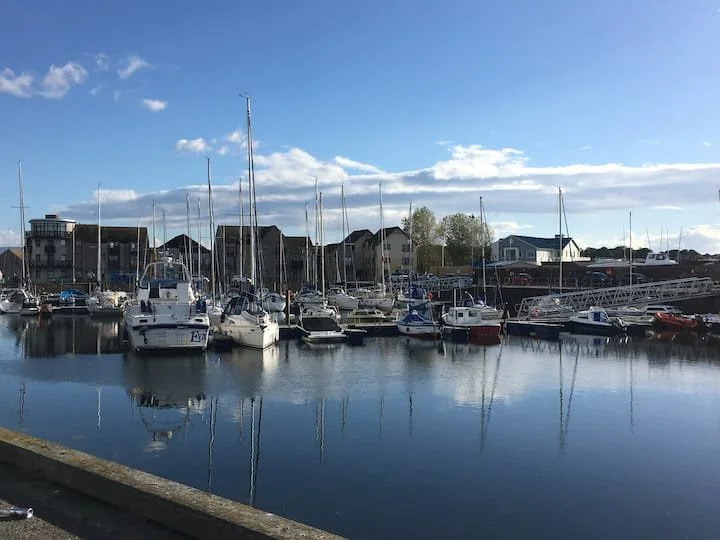 Boats docked at a marina with residential houses in the background under a partly cloudy blue sky.