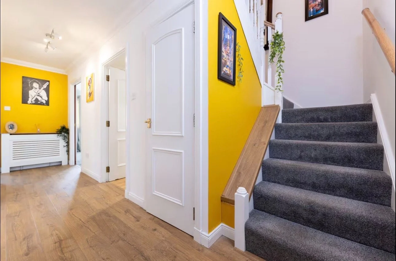 Interior view of a house hallway with wooden flooring, white walls, and a staircase with gray carpet leading upstairs. There is a yellow accent wall with framed artwork and hanging plants. A white door with decorative paneling is partially visible.