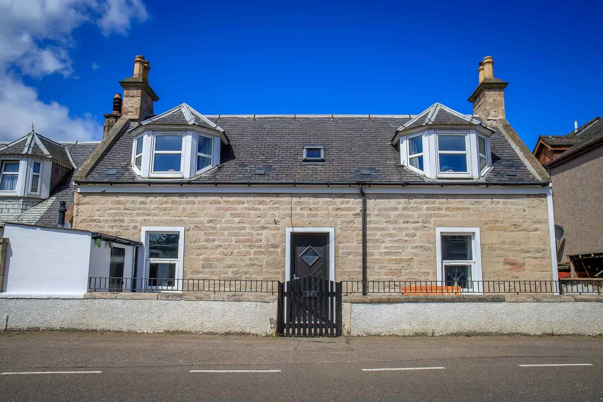 A two-story stone house with a slate roof, white window frames, and a black front door, surrounded by a small wall and metal railing. The house has two large bay windows on the upper floor and one window on each side of the door on the ground floor. 