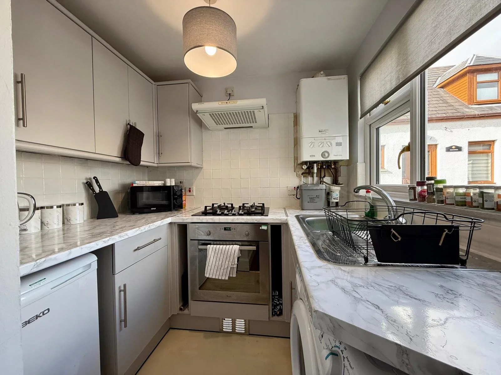A small kitchen with white cabinets, marble countertop, microwave, stove, small oven, and a dish drying rack by a sink near large windows. Outside view shows neighboring buildings.