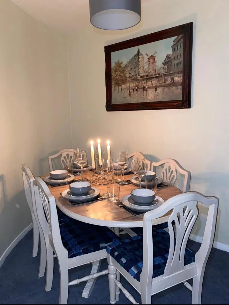 A dining room with an oval table set for six, with white chairs featuring dark blue patterned cushions, a candelabra with lit candles, and a painting of a cityscape hanging on a cream-colored wall.