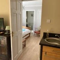 View of a bedroom seen through an open door, with a bed, small nightstand, and a window. Part of a kitchen with a sink and countertop is visible in the foreground.