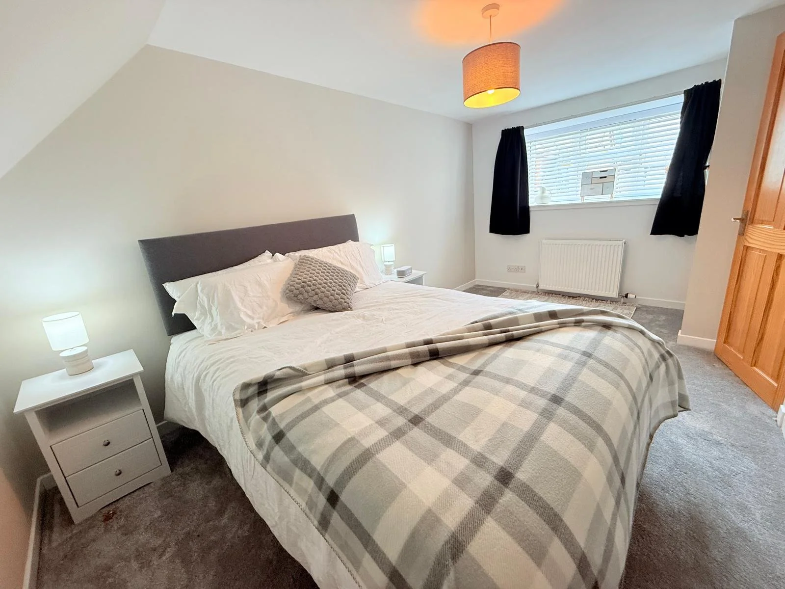 A neatly made bed with white linens and a plaid blanket, two white nightstands with lamps, a window with black curtains, and a wooden wardrobe in a bedroom with light-colored walls and carpeted floor.