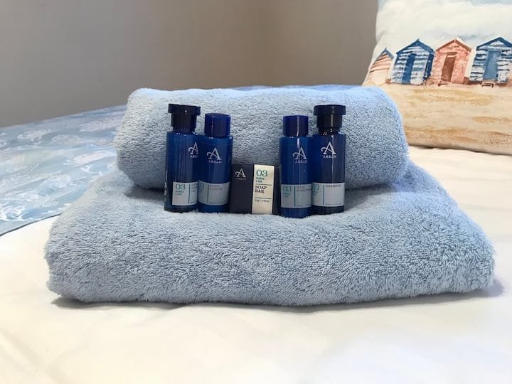 Stack of two light blue towels with four small bottles of toiletries and a soap bar on top, placed on a bed with a decorative pillow in the background.