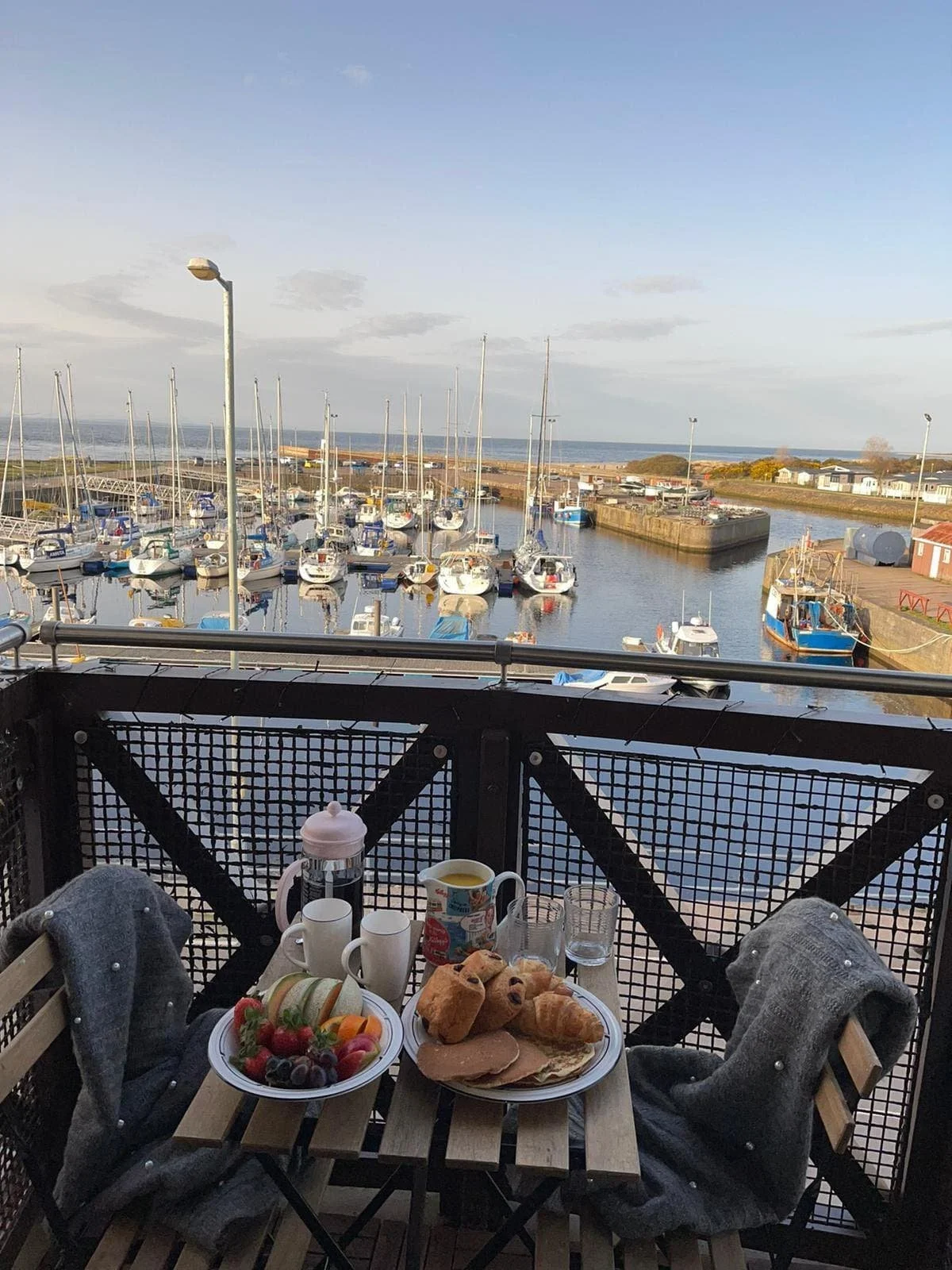 A breakfast table on a balcony overlooking a marina with sailboats and a coastal landscape in the background.
