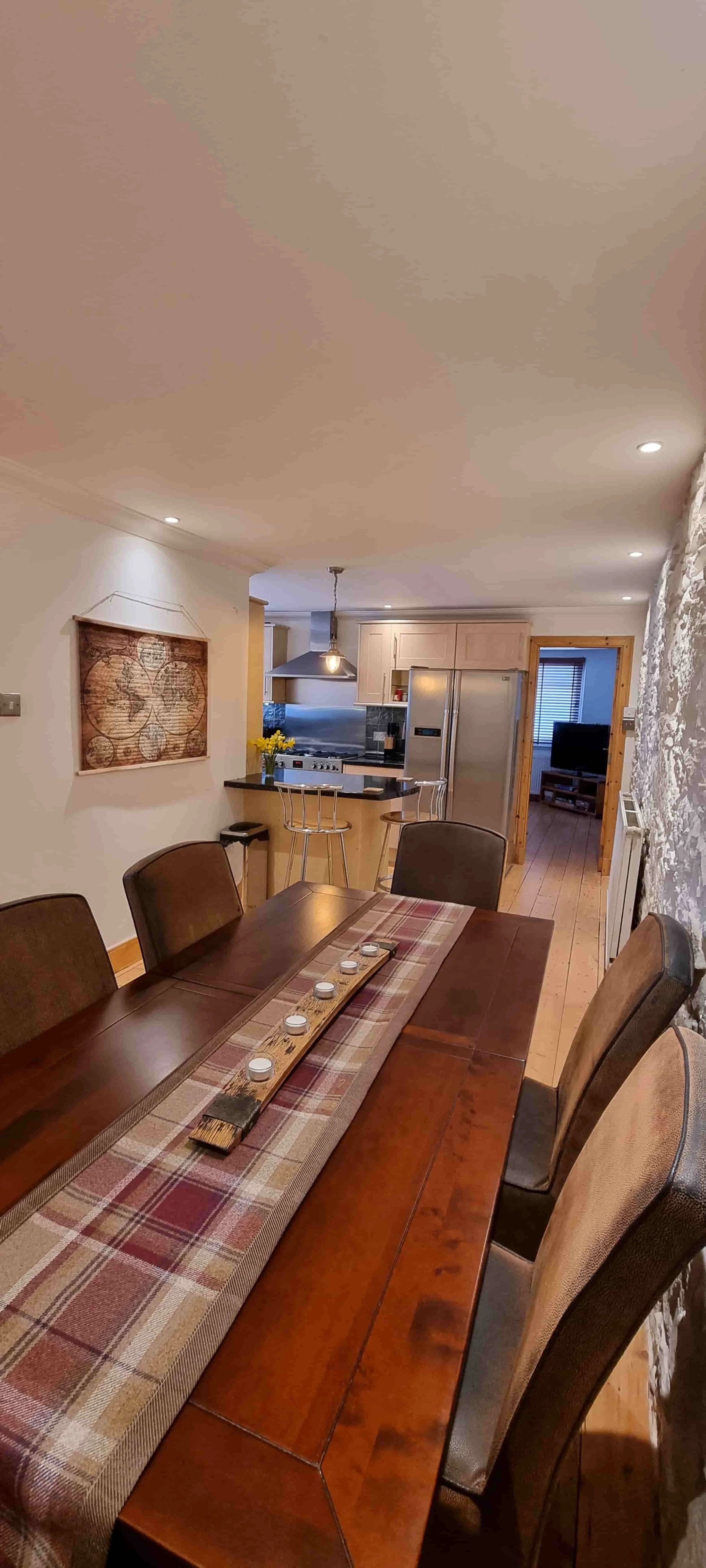 A dining room with a wooden table and six chairs, a plaid table runner with candles, a kitchen area in the background with a small island, six barstools, a refrigerator, and a doorway leading to a living room with a television.