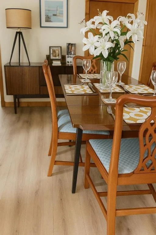 Dining room with a wooden table set for a meal, decorated with a large vase of white lilies, placemats, glasses, and cutlery, with a mid-century modern sideboard, framed pictures, and a lamp in the background.