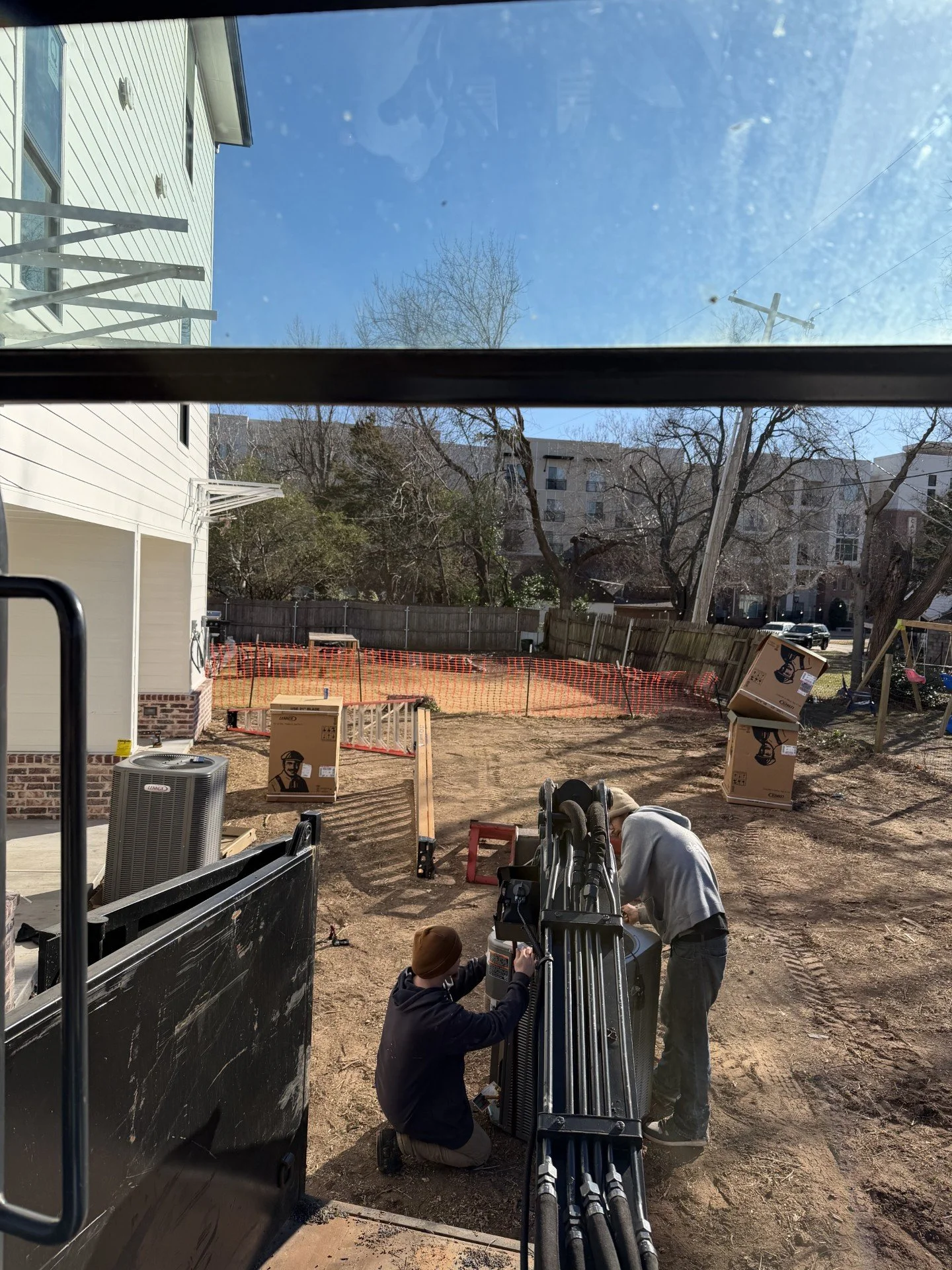 Two workers are installing equipment in a backyard construction site with dirt ground, wooden fences, and equipment boxes, viewed from inside a vehicle.