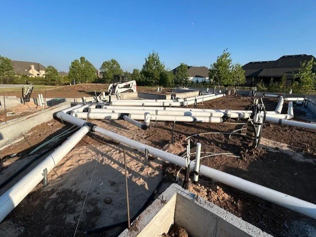 Construction site with large white PVC pipes installed on the ground, and surrounding houses and trees under a clear blue sky.