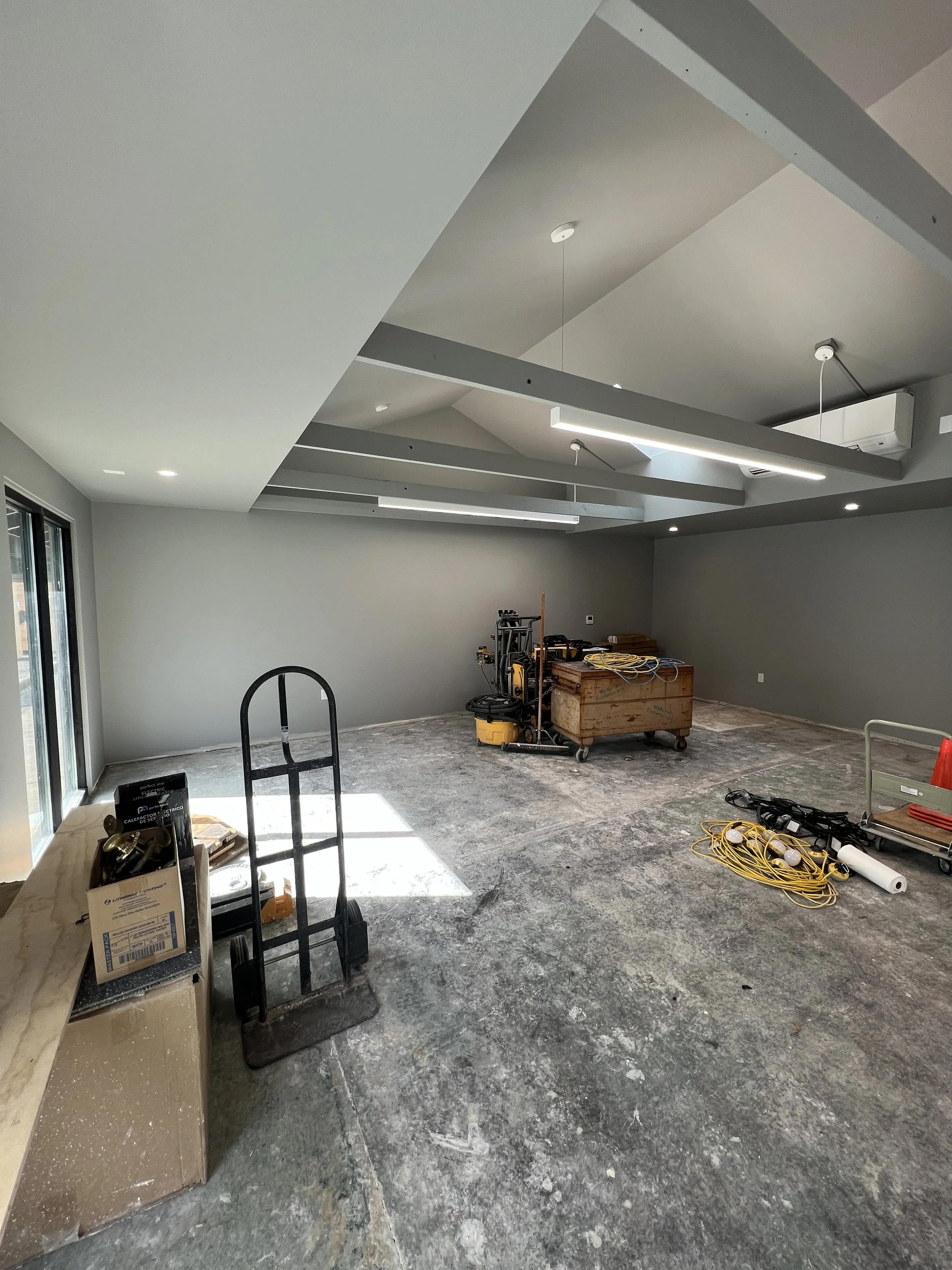 Interior of a room under construction with gray walls and ceiling, construction tools, cords, and equipment scattered on the floor, and large sliding glass doors on the left side.