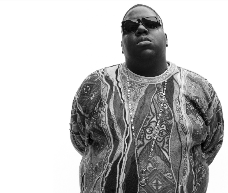 Biggie Smalls Notorious B.I.G. wearing black sunglasses, a coogi sweater and a chain necklace against a plain background.