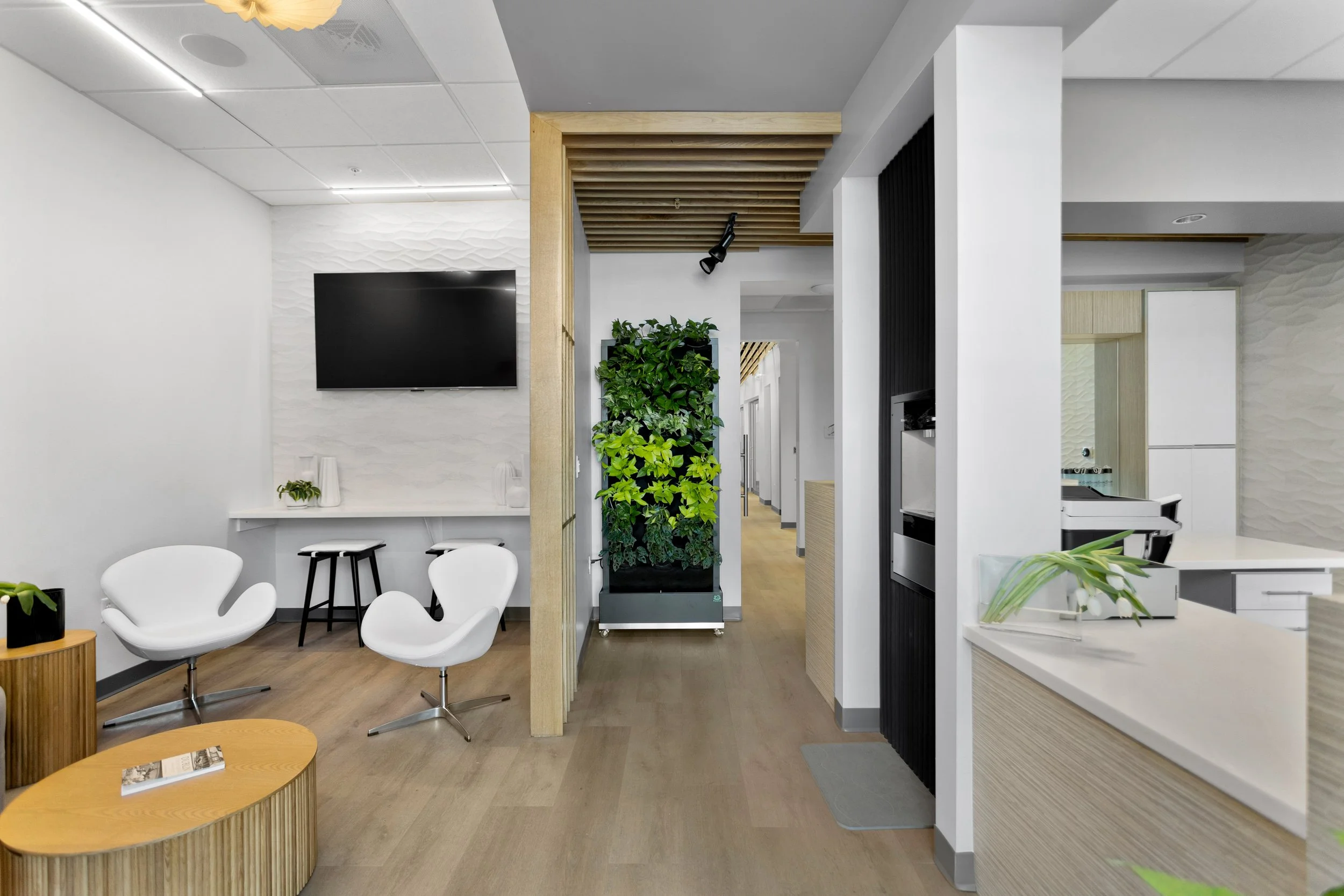 Patient work desk with stools, tulip chairs and japandi style tables. A living wall is shown beneath acoustic wood columns and beams. Goodbye mirror, flat screen t.v., water filler.