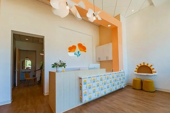 Interior of a modern reception area with a colorful, patterned counter, a white wall with orange abstract art, a small seating area with yellow stools, and wooden flooring.