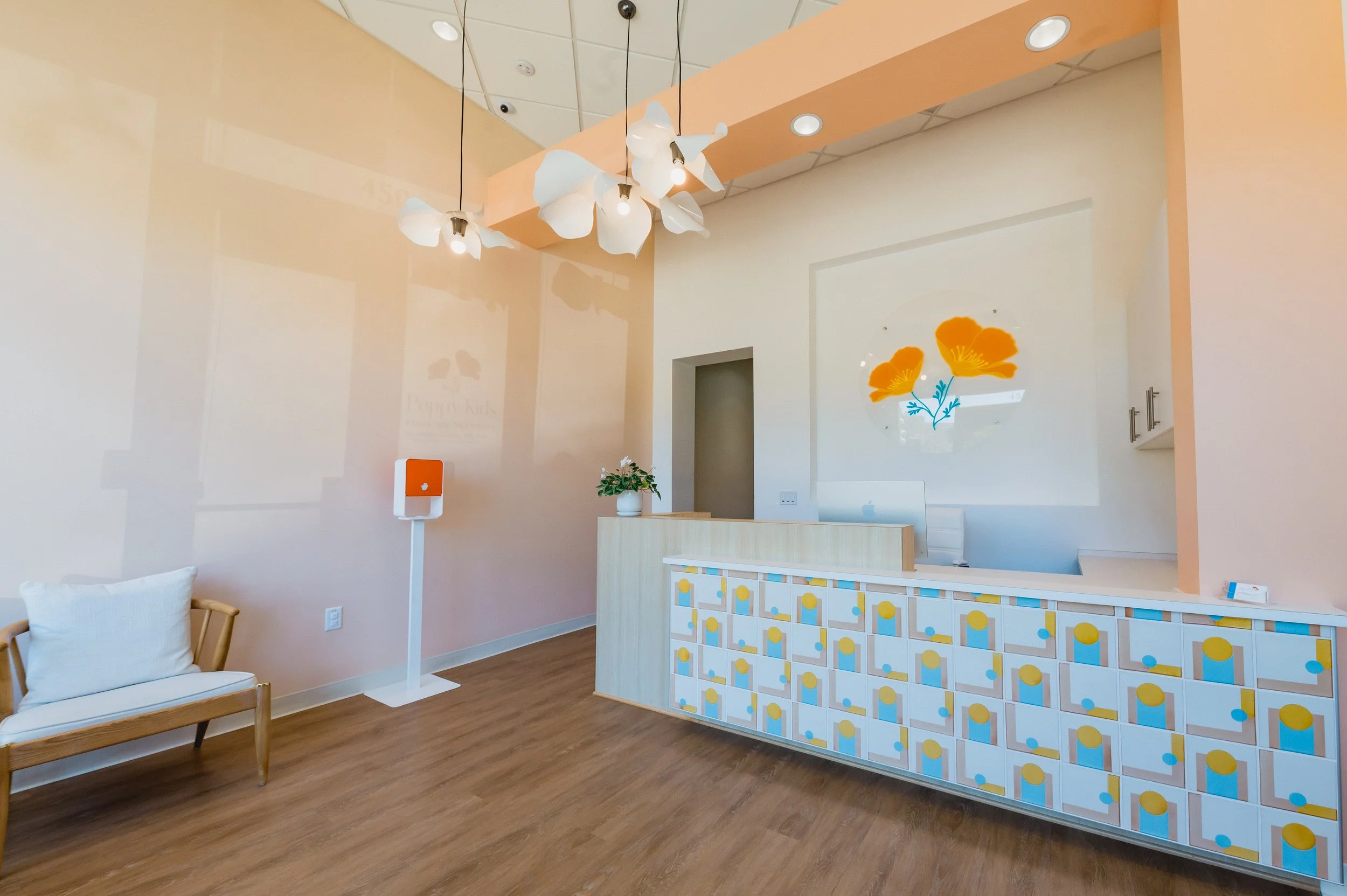 Modern reception area with pastel-colored decor, a patterned front desk, a white bench with a pillow, and a wall art of orange flowers. Contains a flower vase, a small white and orange stand, and ceiling lighting.