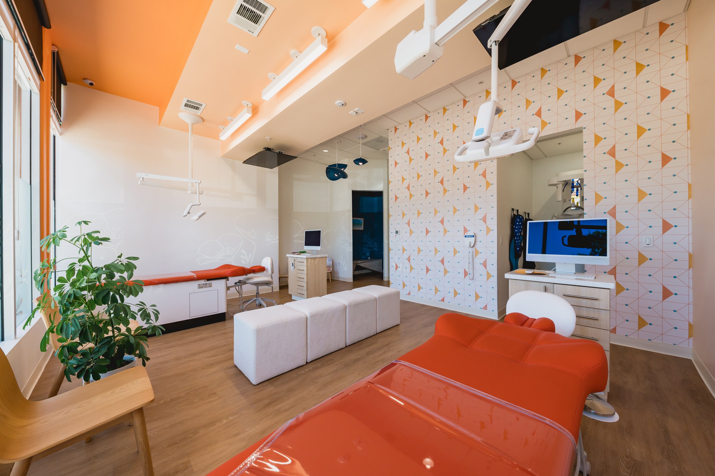 A modern dental clinic room with orange and white decor, dental equipment, a dental chair, a computer, and seating area.