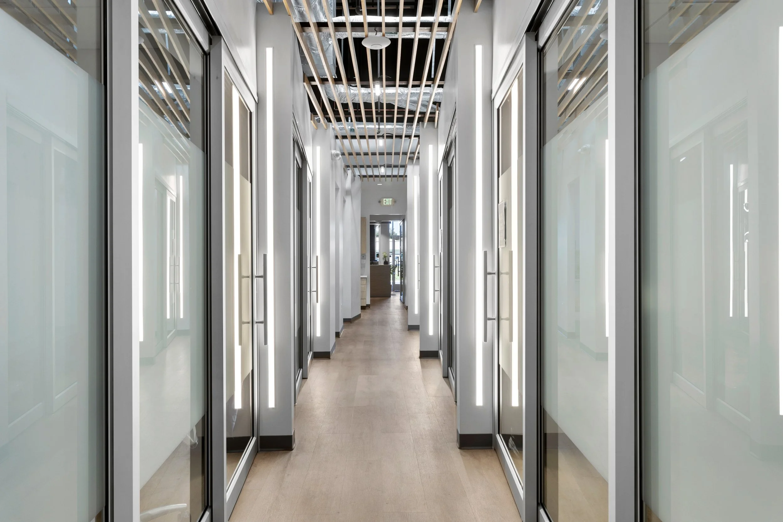Angled hallway with acoustic beam ceilings, glass walls and doors and vertical lighting