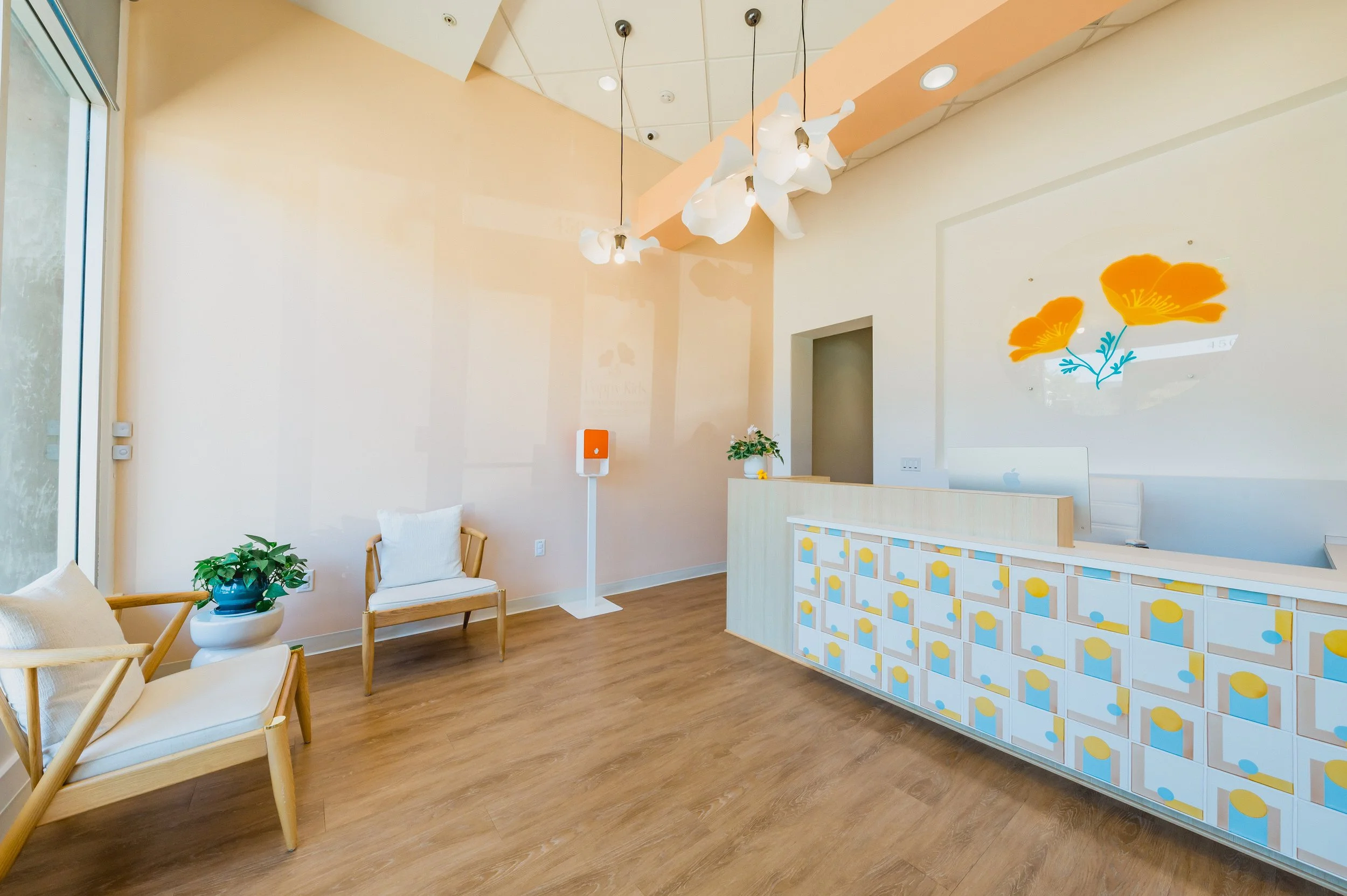 Modern reception area with wooden flooring, a decorated front desk, and seating with white cushions. There are potted plants, artistic ceiling lights, and a floral wall art.
